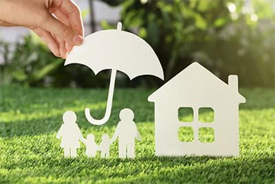 Une femme tenant un parapluie en papier protégeant une famille posée sur l'herbe à côté d'une maison en papier