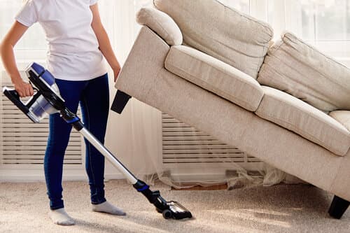 Une personne habillée avec un-shirt blanc et un jean, soulevant le canapé pour passer l'aspirateur