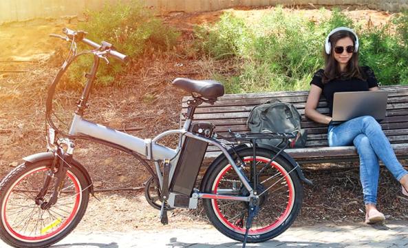 Un vélo électrique posé devant un banc sur lequel est assise une jeune femme avec son ordinateur portable