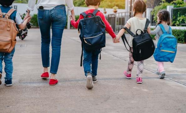 Des enfants avec des cartables et une femme de dos se tenant tous par la main