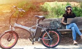 Un vélo électrique posé devant un banc sur lequel est assise une jeune femme avec son ordinateur portable