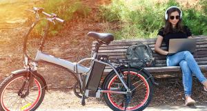 Un vélo électrique posé devant un banc sur lequel est assise une jeune femme avec son ordinateur portable