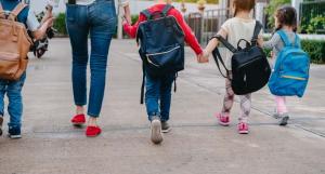 Des enfants avec des cartables et une femme de dos se tenant tous par la main
